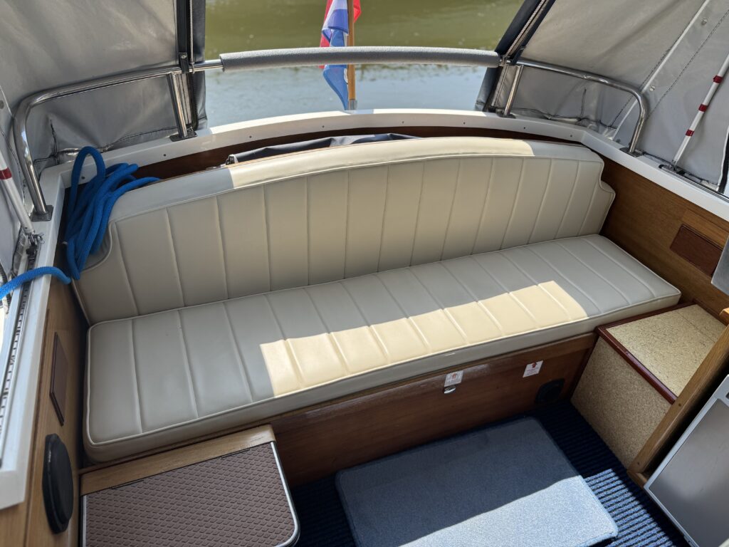 A spacious, light-colored upholstered bench seat on a boat, positioned against a wooden backrest. The interior includes a small table to the left, a blue mat on the floor, and a view of water and a flag through a window.
