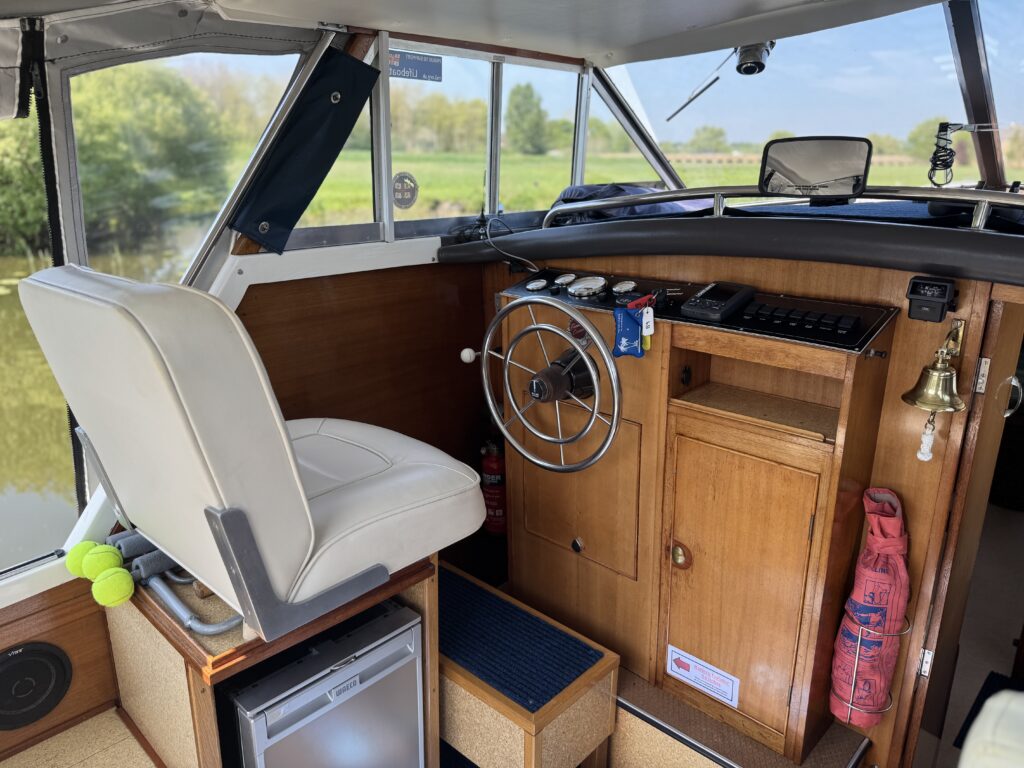 The interior of a boat's control area featuring a white captain's chair beside a wooden steering wheel. There are various navigation instruments on the dashboard, a fire extinguisher, and a bell mounted on the side. Large windows offer views of greenery outside.