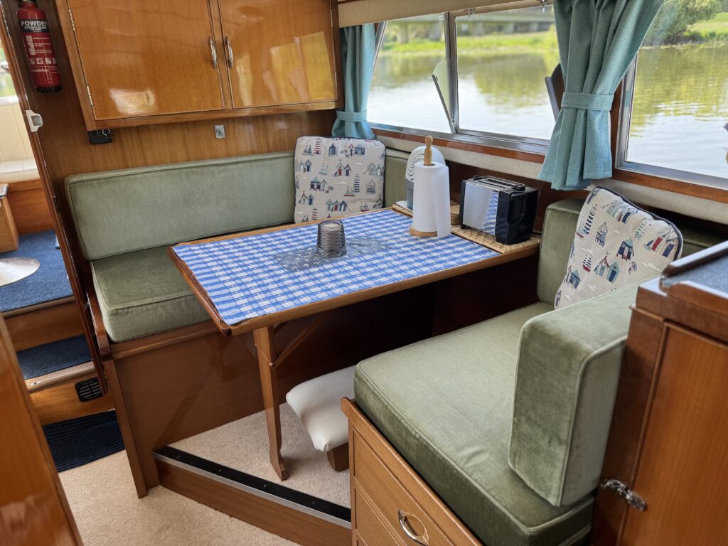 An interior cabin space featuring a small dining table with a blue and white checkered tablecloth. Surrounding the table are cushioned benches with floral-patterned upholstery. Windows provide a view of greenery outside, and a small appliance is visible on the table.