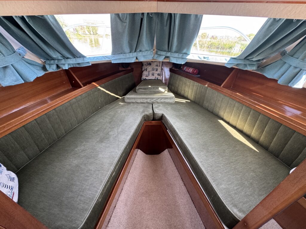 A cabin interior of a boat featuring two green upholstered seating areas arranged in a V-shape. Light-colored wooden panels and curtains decorate the space, and a single cushion is placed at the center. The sunlight illuminates the area.