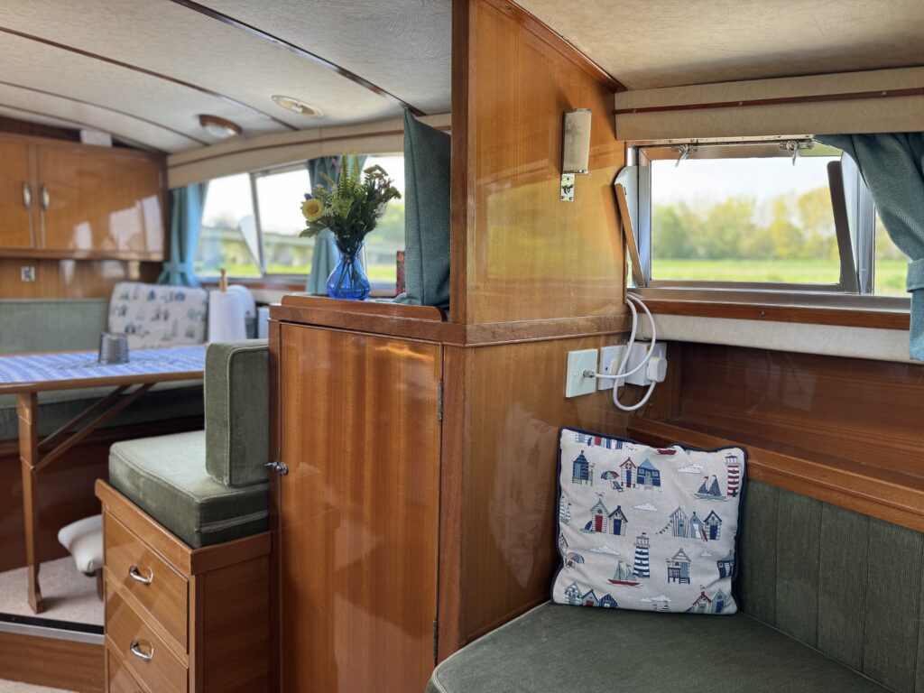 An interior view of a boat featuring wooden paneling, a green upholstered seat, and a white cushion with a nautical design. A table with a blue tablecloth and a vase of yellow flowers is visible in the background, along with large windows letting in natural light.