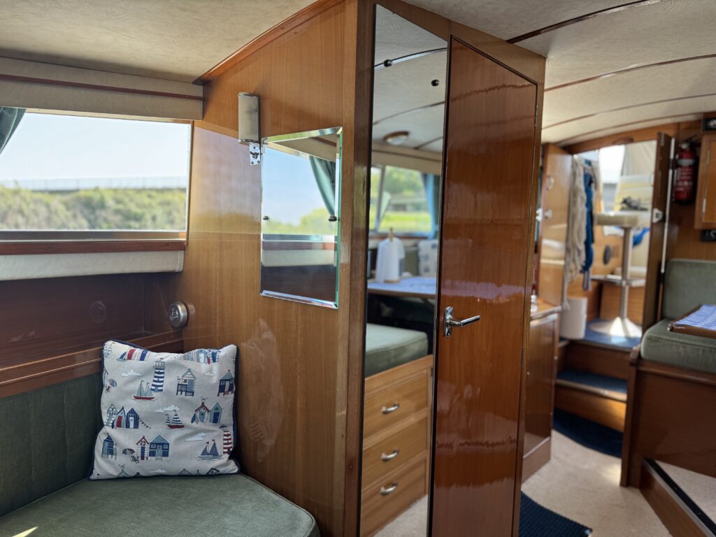 The interior of a boat features wooden paneling and a green cushioned seating area with a decorative pillow. A mirror is mounted on the wall, and a door leads to another section of the boat. Sunlight streams in through windows, illuminating the space.
