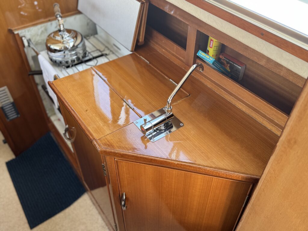 A wooden counter with a polished surface features a stainless steel sink faucet on the right side. To the left, there is a gas stovetop with a pot, and a small cabinet below contains an organized space. The background includes light-colored walls and a dark rug.