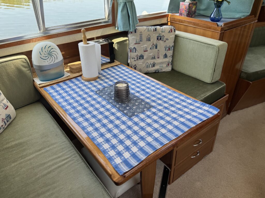 A small dining table on a boat covered with a blue and white checkered tablecloth. Centered on the table is a lantern and a paper towel holder, alongside a decorative fan. Green upholstered seating is visible in the background, creating a cozy atmosphere.