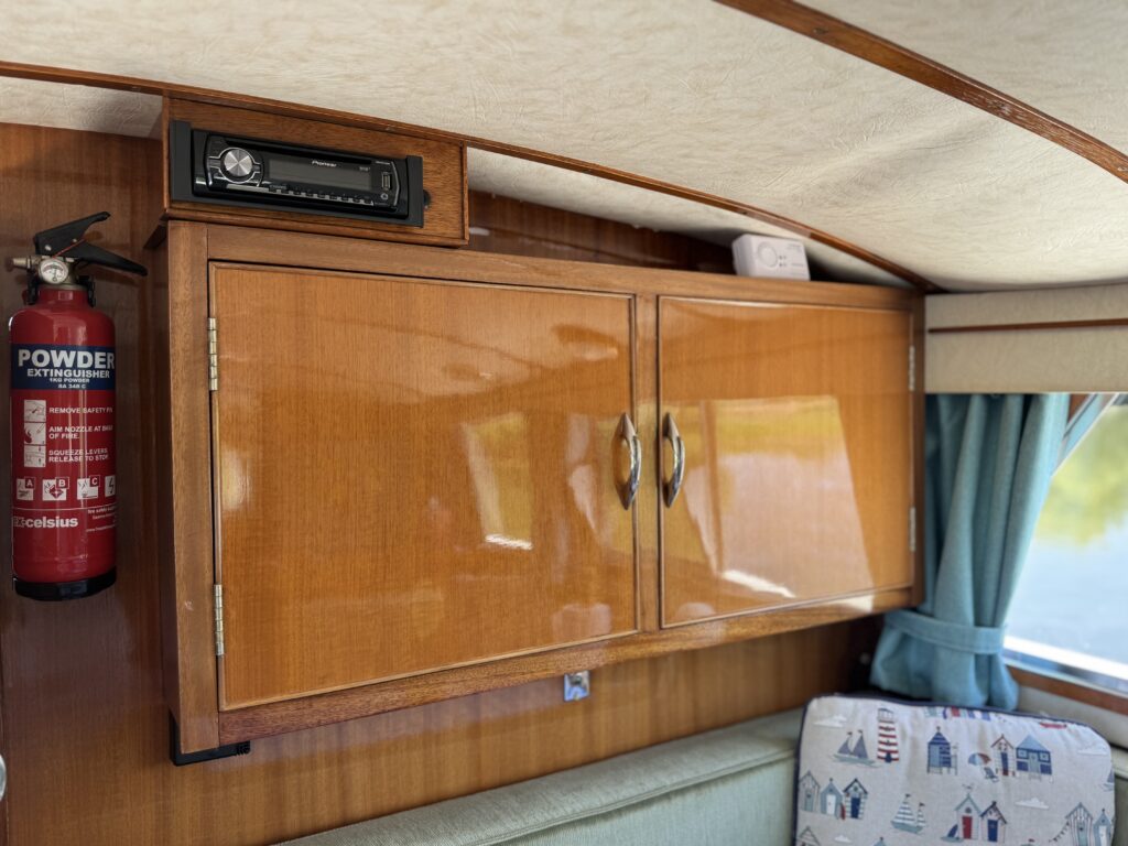 Interior of a boat featuring a wooden cabinet with two doors, a radio mounted above, and a red fire extinguisher to the left. A light green sofa with decorative fabric is visible in the foreground, and blue curtains frame a window with a view outside.