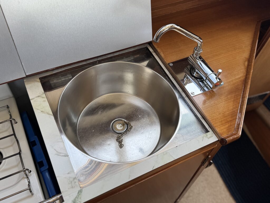 A stainless steel sink with a circular shape is installed in a wooden countertop. A faucet is positioned to the right of the sink, and the surrounding area features a marble trim. Nearby, there is a gas stove and part of the kitchen cabinetry.