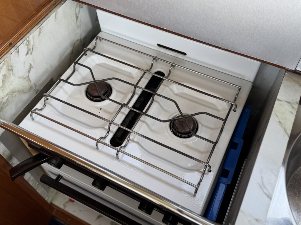 A white two-burner gas stove is installed in a kitchen cabinet. The burners are covered with metal grates, and the stove is surrounded by a gray countertop and wooden cabinetry.