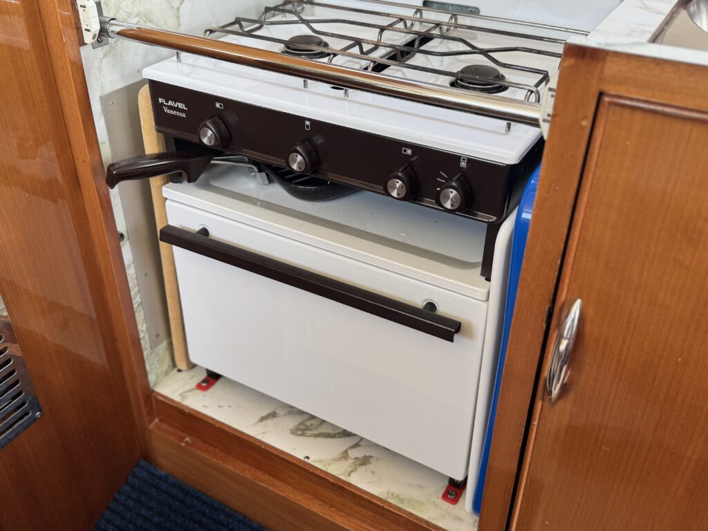 A compact stove and oven combination installed in a wooden kitchen cabinet. The stove has two burners and four control knobs on the front, while the oven below features a white door with a flat handle and is situated above a blue object.