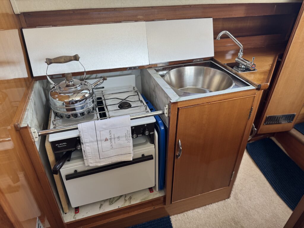 A compact kitchen area featuring a silver kettle on a stovetop next to a built-in sink. Below the counter, there is a refrigerator with a drawer, and wooden cabinetry surrounds the space. The flooring is covered with a light-colored carpet.