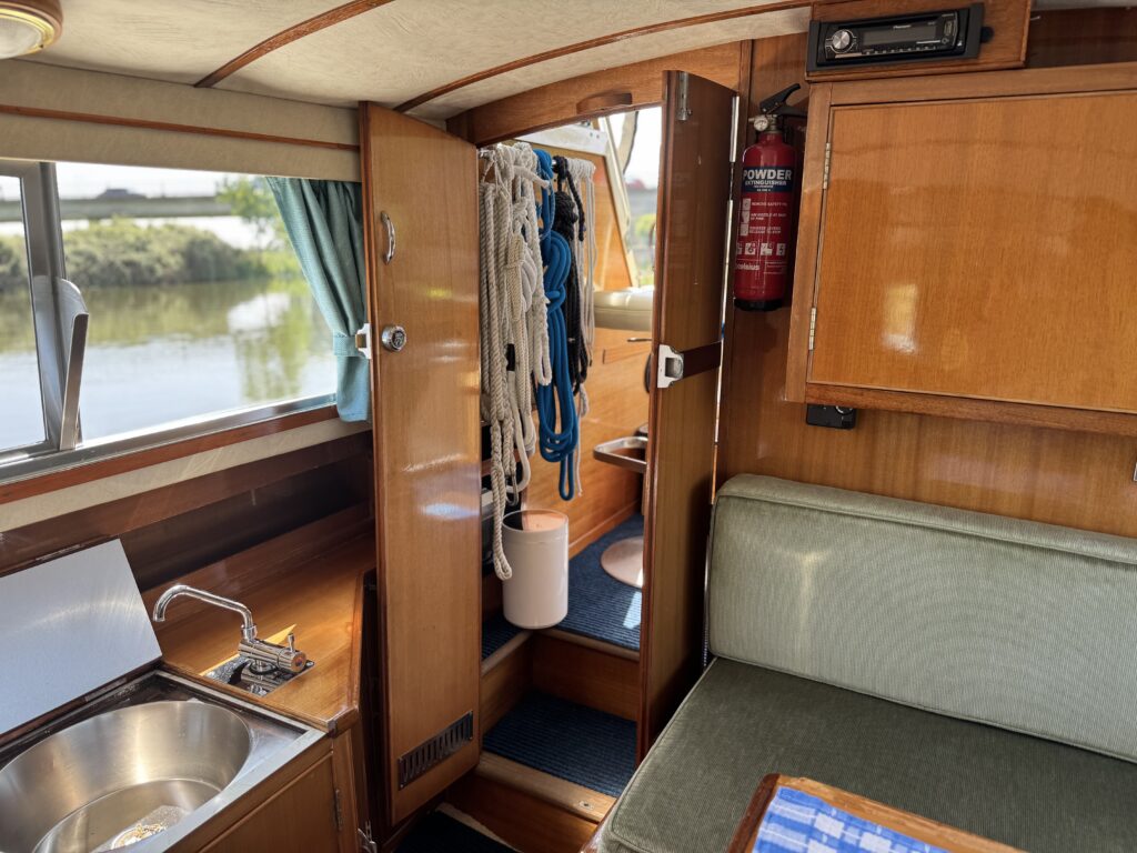 The interior of a boat features polished wooden cabinetry, a small sink, and a green upholstered bench. Ropes hang on the wall near a door leading to a space with visible plumbing, and large windows offer a view of greenery outside.