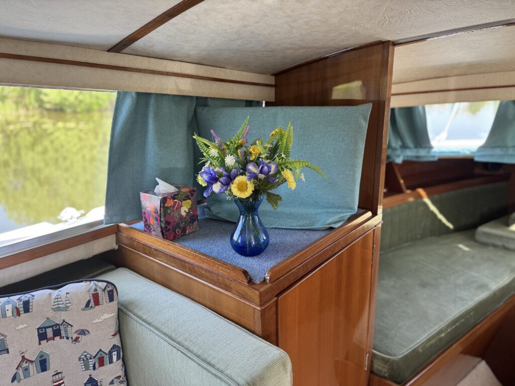 A vibrant bouquet of yellow and purple flowers arranged in a blue glass vase sits on a wooden surface inside a boat cabin. In the background, a teal fabric panel and a large window reveal a view of greenery and water outside.