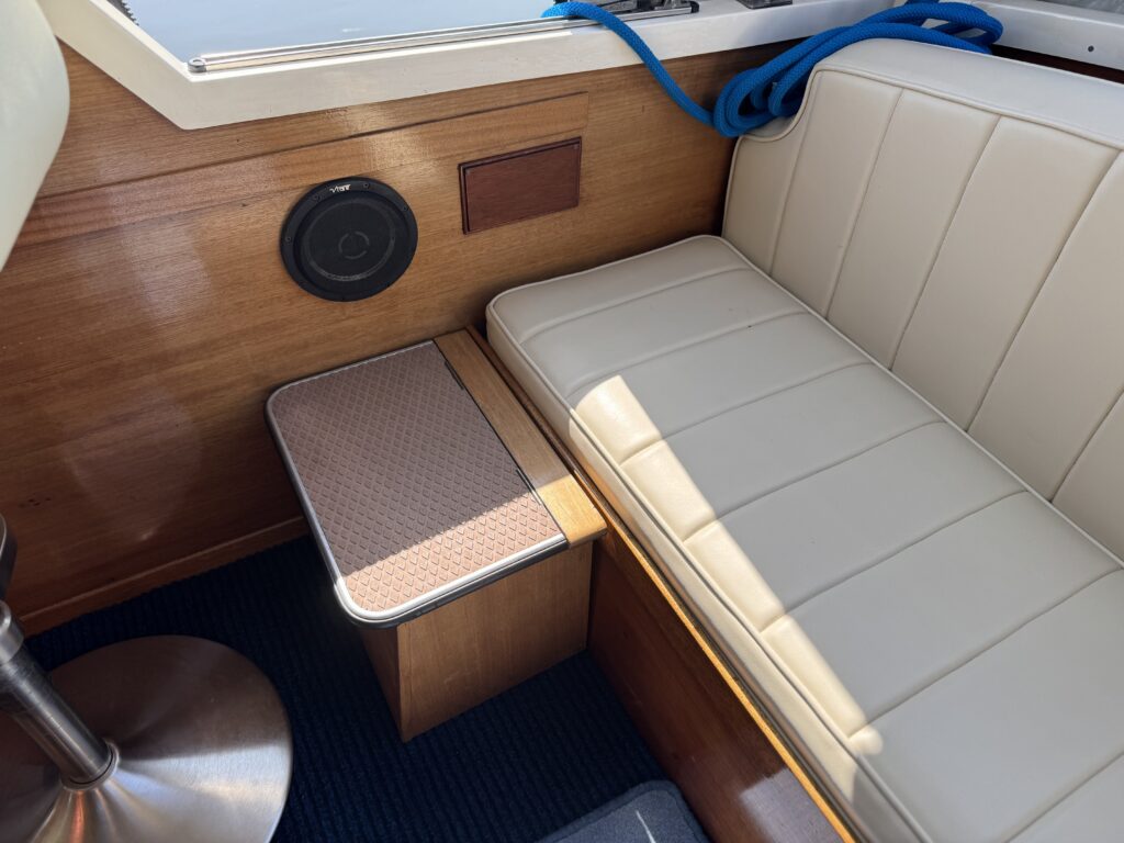 A light-colored leather seat cushion is positioned beside a wooden table on a boat. The table has a textured surface and a round speaker is mounted on the wooden wall in the background. A blue rope is draped over the top edge.