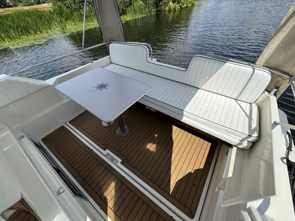 The image shows the interior of a Fairline Sunfury boat. It features a white cushioned seating area with a small table at the center, all set on wooden flooring. The boat's side offers a view of green foliage and water.