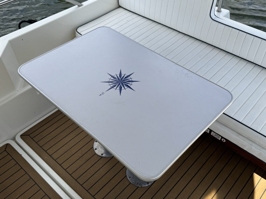 A white table with a blue compass rose design sits on a wooden deck of a boat. The table is supported by two round bases, and the surrounding area features white cushioned seating and a view of water.