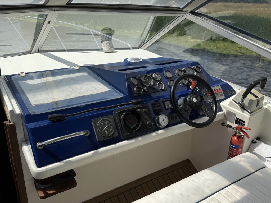 The cockpit of a Fairline Sunfury boat features a blue dashboard with various gauges and a steering wheel. A soft cushioned seat is visible beside a white panel, while a fire extinguisher is mounted nearby.