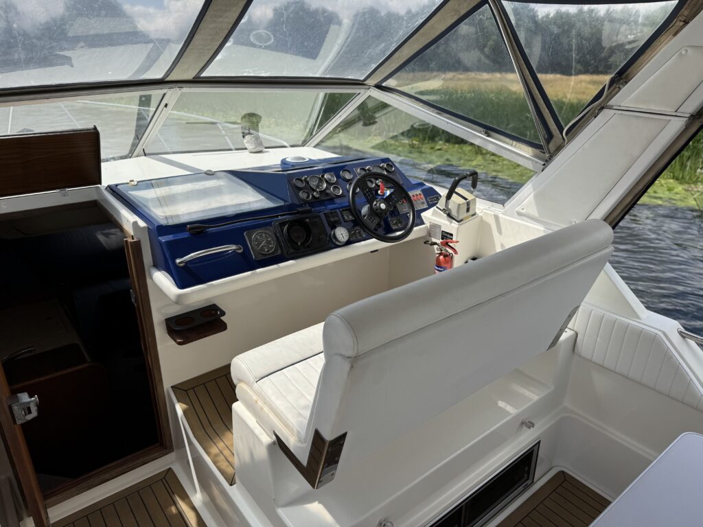 The interior of a Fairline Sunfury boat featuring a white captain's chair next to a blue control panel with various dials and switches. Large windows provide visibility to the water and greenery outside, while wooden flooring adds a nautical touch.