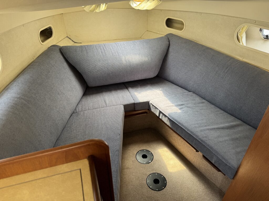 An interior seating area of a Fairline Sunfury yacht, featuring a U-shaped gray upholstered couch. Two round speakers are mounted on the floor, and there are windows allowing natural light to filter in.