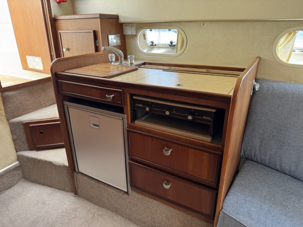 A wooden furniture unit featuring a countertop, a small stainless steel refrigerator, and three drawers. The unit is positioned against a wall with windows nearby, providing natural light to the space.
