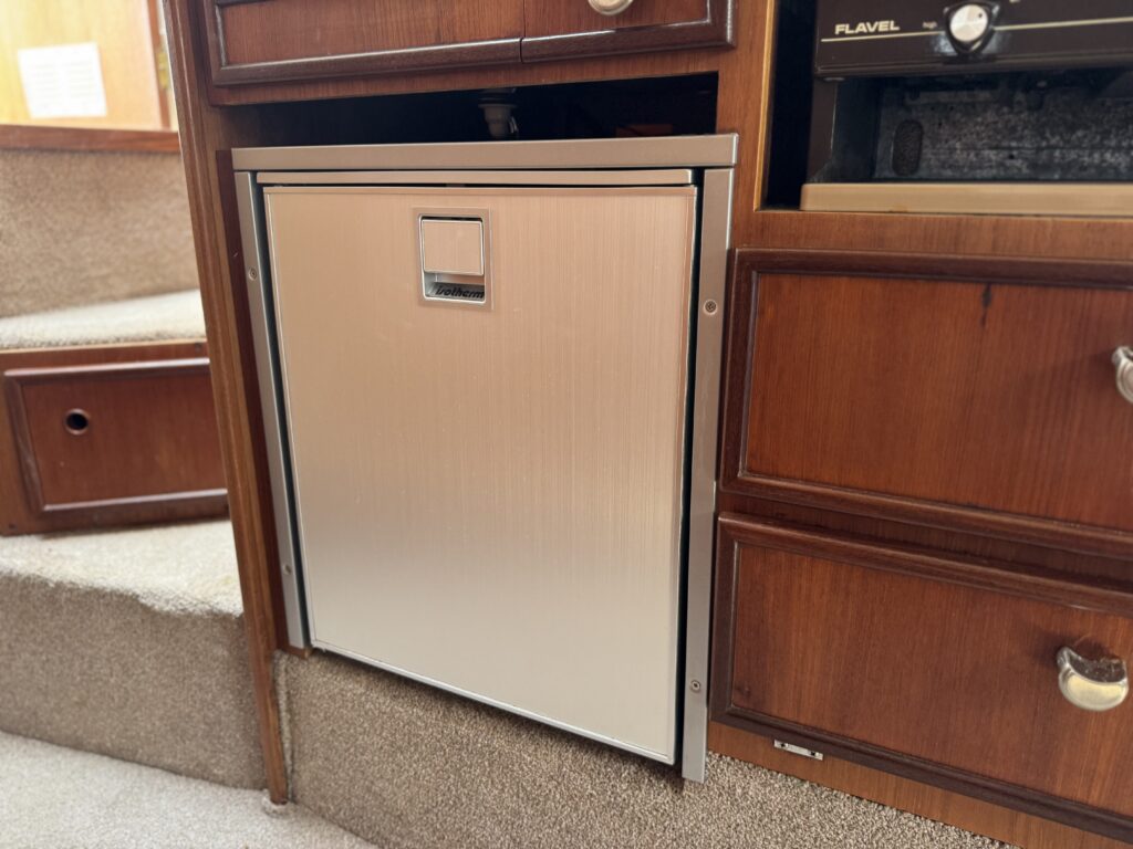A silver metal refrigerator integrated into wooden cabinetry on a boat. The fridge has a rectangular design with a handle and is positioned near a staircase, surrounded by additional wooden storage compartments.