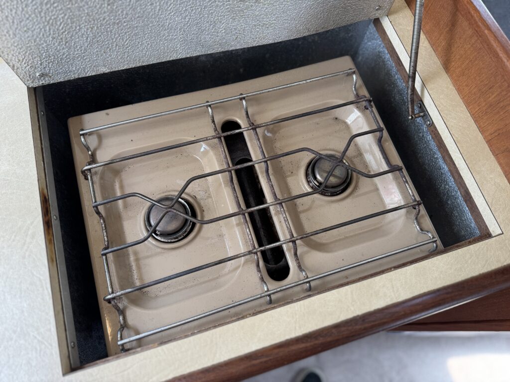 A compact two-burner gas stove with a stainless steel safety grate, set in a wooden cabinet. The stove has a beige enamel surface and is partially covered by a lid.