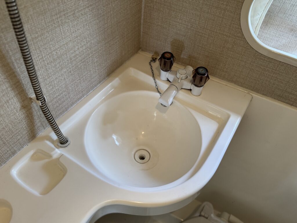 A small circular sink with a smooth white surface, featuring a single faucet and two hot and cold water knobs. The background includes a light-colored textured wall and a window allowing natural light.