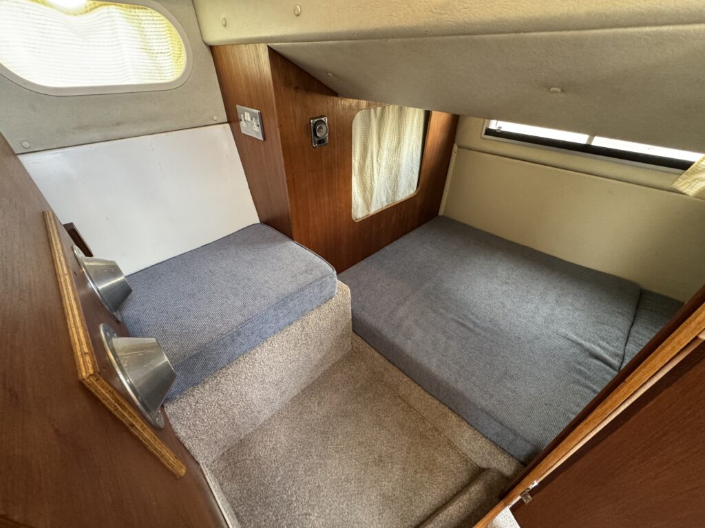 Interior view of a boat cabin featuring a cushioned seating area with two rectangular beds. The space includes light-colored walls, a window with light filtering through, and wood paneling. Stair steps lead to the sleeping area.
