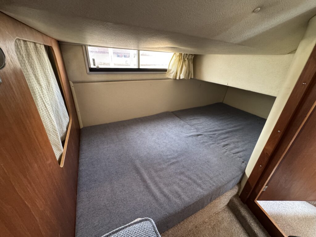 An interior cabin of a Fairline Sunfury boat featuring a large bed with a gray blanket. The walls are upholstered in light-colored fabric, and there is a window with pale curtains allowing natural light to enter.