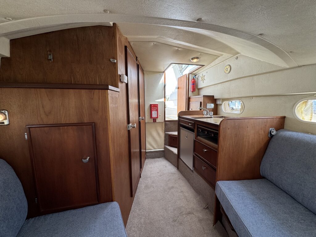 The interior of a Fairline Sunfury boat shows wooden cabinets and a small kitchenette with a sink and drawers. Two blue upholstered couches are positioned along the walls, and windows allow natural light into the cabin.
