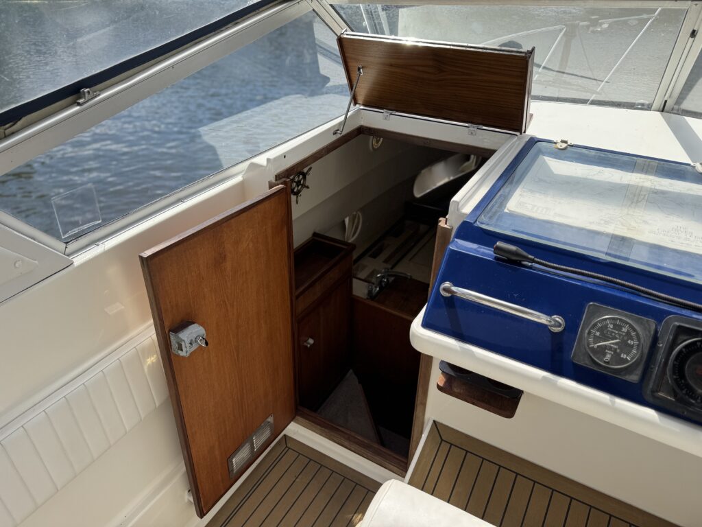 A view of the interior of a Fairline Sunfury boat, showing an open wooden door leading to a small cabin below deck. The area includes wooden cabinetry and a control panel with gauges on a countertop. Sunlight streams in through a nearby window.