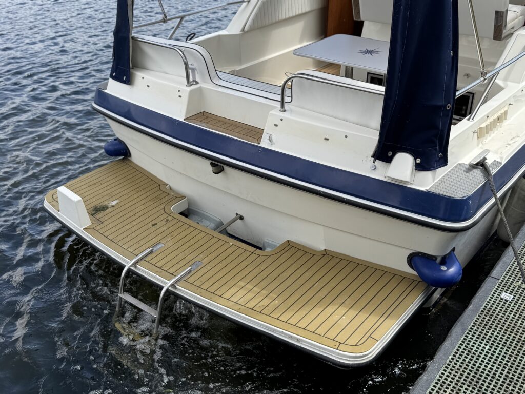 The rear view of a Fairline Sunfury boat shows its beige synthetic teak deck and a ladder leading into the water. The boat has a blue and white color scheme with a canopy above the deck area. The surface reflects sunlight on the water.