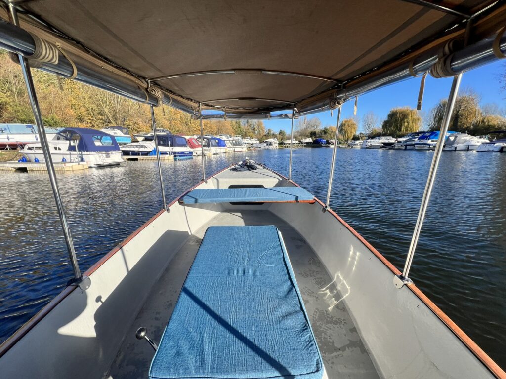 The interior of a boat showing a central blue mat on the deck. The boat is surrounded by water and moored boats are visible in the background under a clear blue sky with some trees nearby.