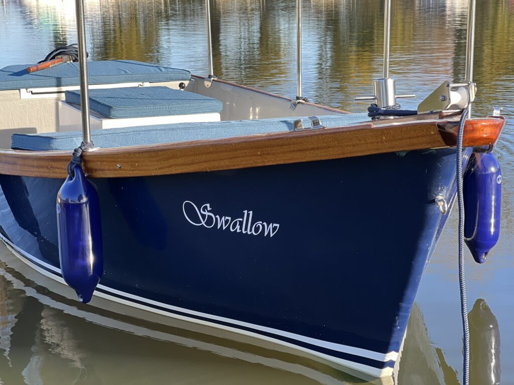 A blue boat with a wooden trim is docked, featuring the name "Swallow" painted in white on the bow. Two blue fenders hang from the sides, reflecting in the calm water.