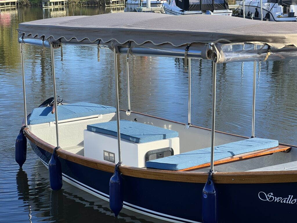 A blue and white boat with a canopy sits on calm water, featuring two blue cushions on the seat and a white compartment in the center. The boat has blue fenders hanging over the side and is surrounded by other boats in the background.