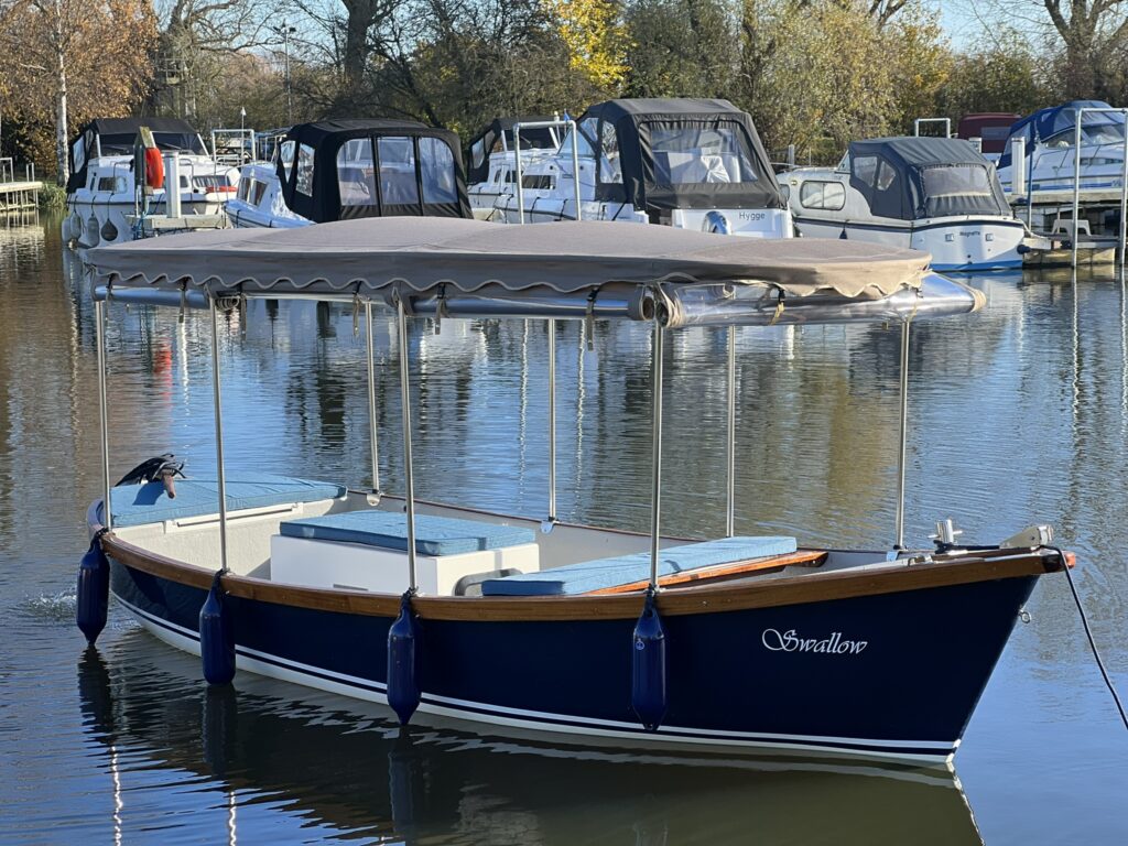 A blue and white boat named "Swallow" with a canopy docked in calm water. Several other boats with covers are visible in the background, surrounded by trees with autumn foliage.