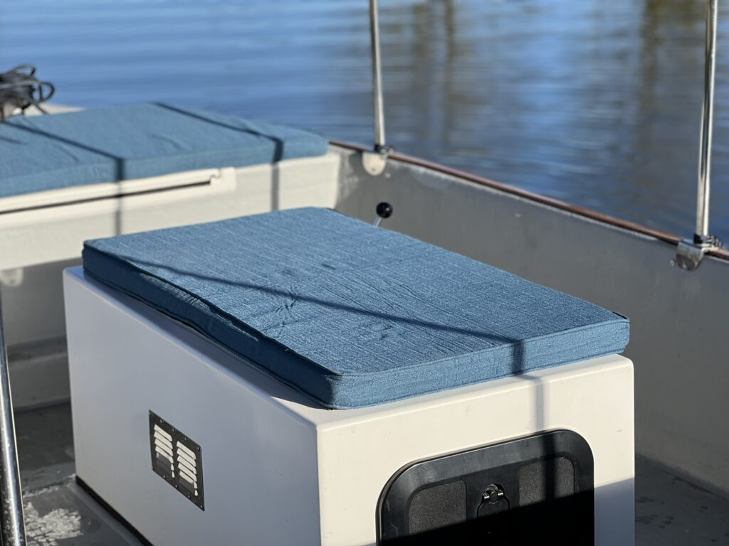A blue cushion rests on a white storage box in a boat. The background features reflective water and the sunlight creating a calm setting with no additional items visible in the immediate area.