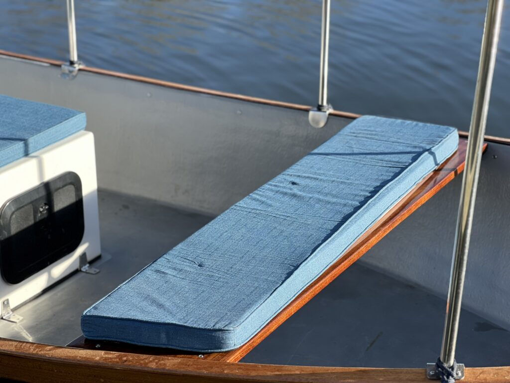 A blue padded bench cushion placed on a wooden boat near the side, with a white storage compartment visible in the background. The surface of the water reflects sunlight, creating a serene outdoor setting.
