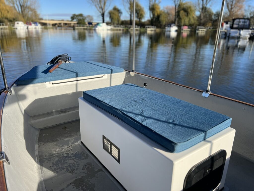 The interior of a small boat features a white storage compartment with a blue cushion on top, and a matching blue cushion on the left side. The calm water and trees in the background reflect the peaceful surroundings.