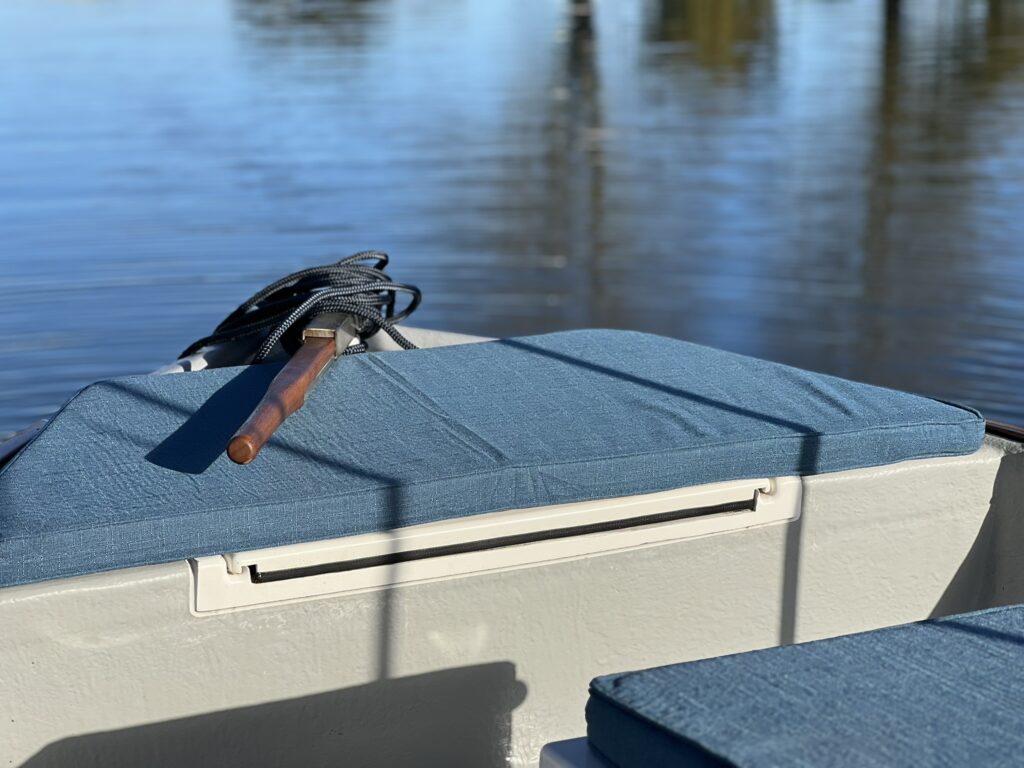 The interior of a small boat featuring blue cushions on the seats. A coiled rope lies next to a wooden handle on a box, with the water surface reflecting trees and sunlight in the background.