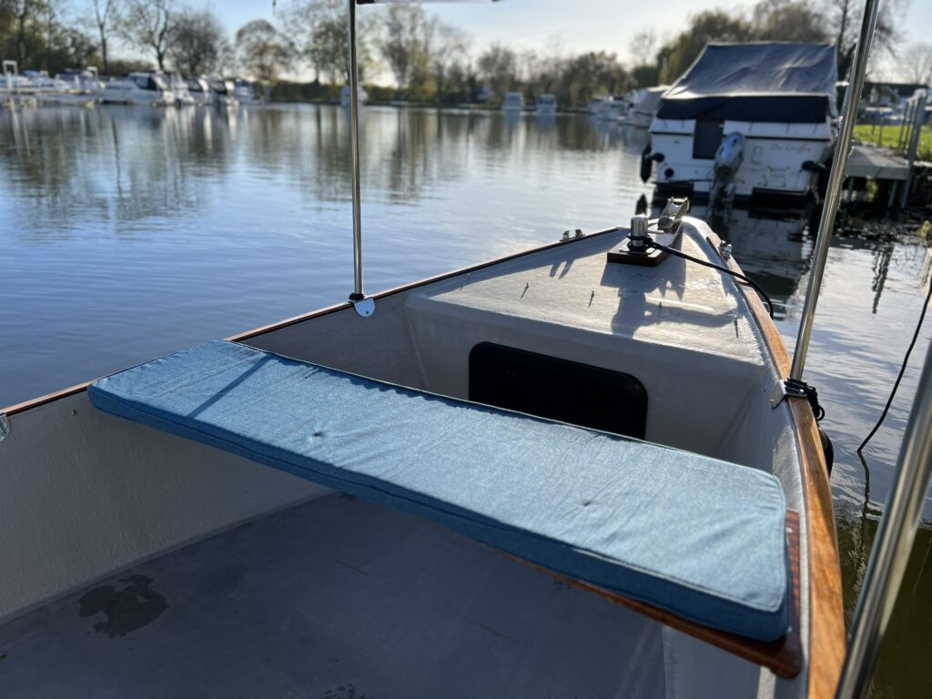 The interior of a boat shows a blue cushion on the bench seat with a smooth wooden edge. The scene includes still water reflecting trees and moored boats under a clear sky. Sunlight illuminates the area creating a calm atmosphere.