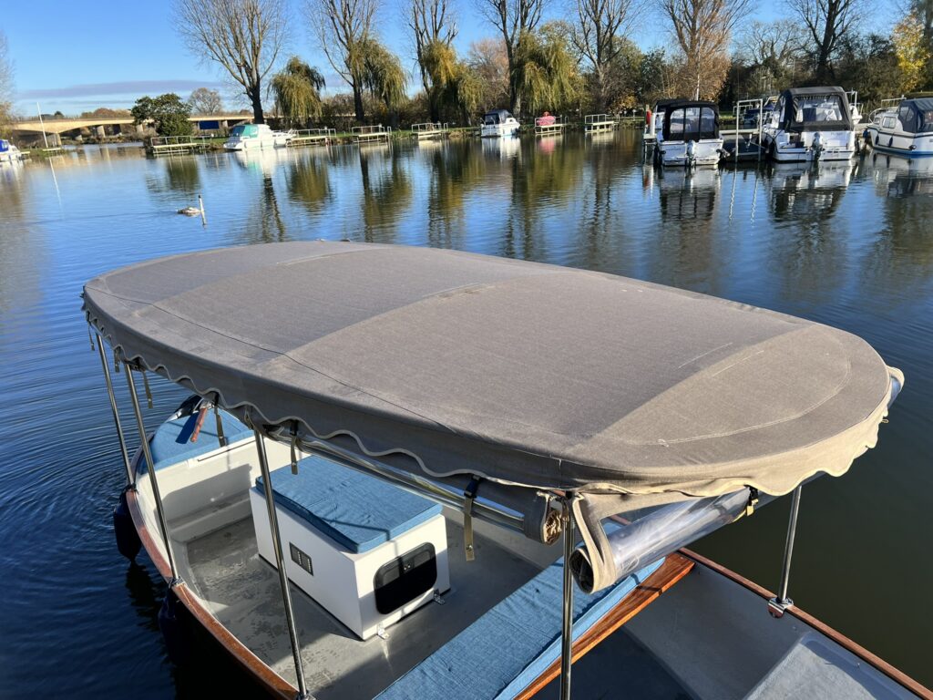 A boat with a brown canopy is moored on calm water, reflecting surrounding trees. The boat features light blue seats and a gray interior, with other boats visible in the background.