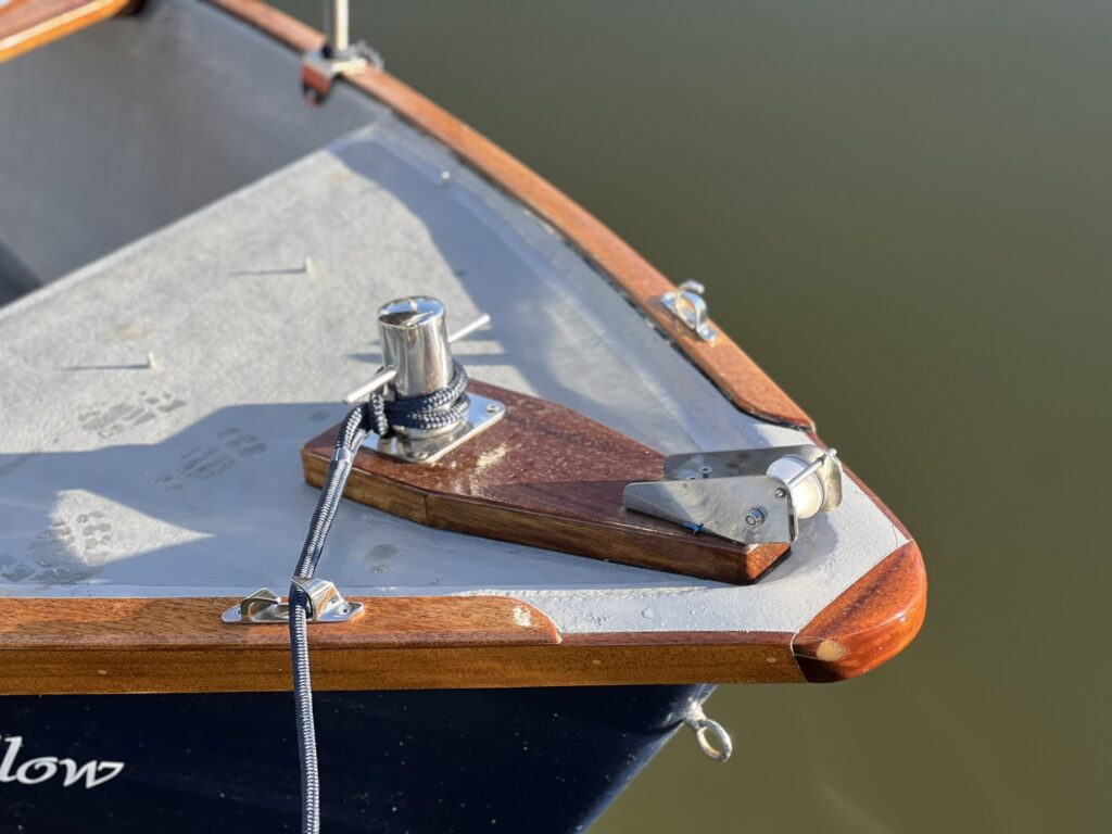 The close-up view of a wooden boat's bow featuring a polished wooden mount for a cleat and a metal fairlead. The boat's deck is gray, with a few markings visible, contrasting against the deep blue hull and calm water in the background.