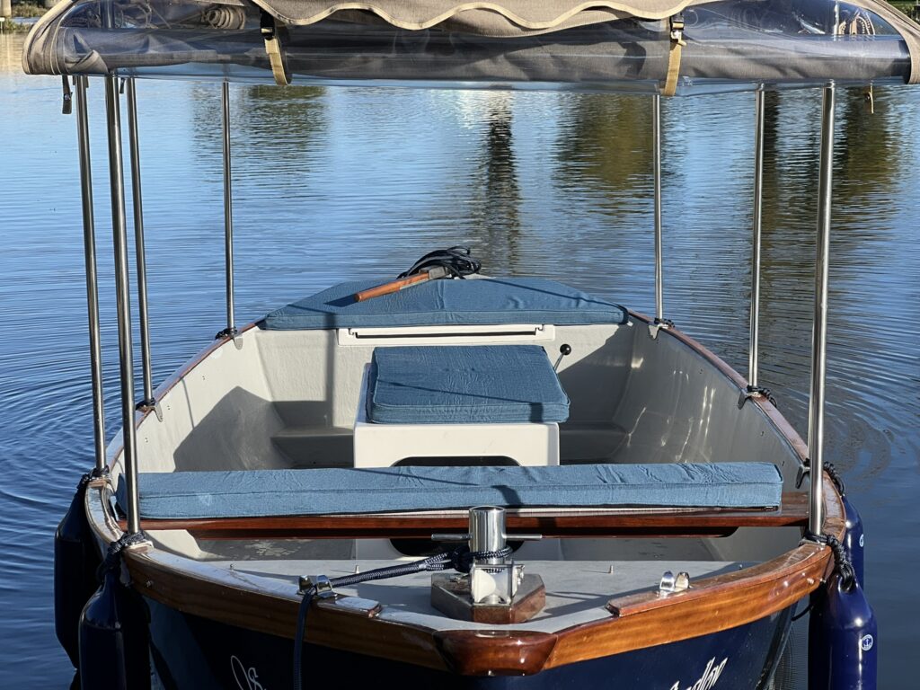 A view of a small boat anchored on calm water. The boat features blue cushions on the seats, a wooden interior, and a canopy above. The surrounding area reflects in the water, creating a serene atmosphere.