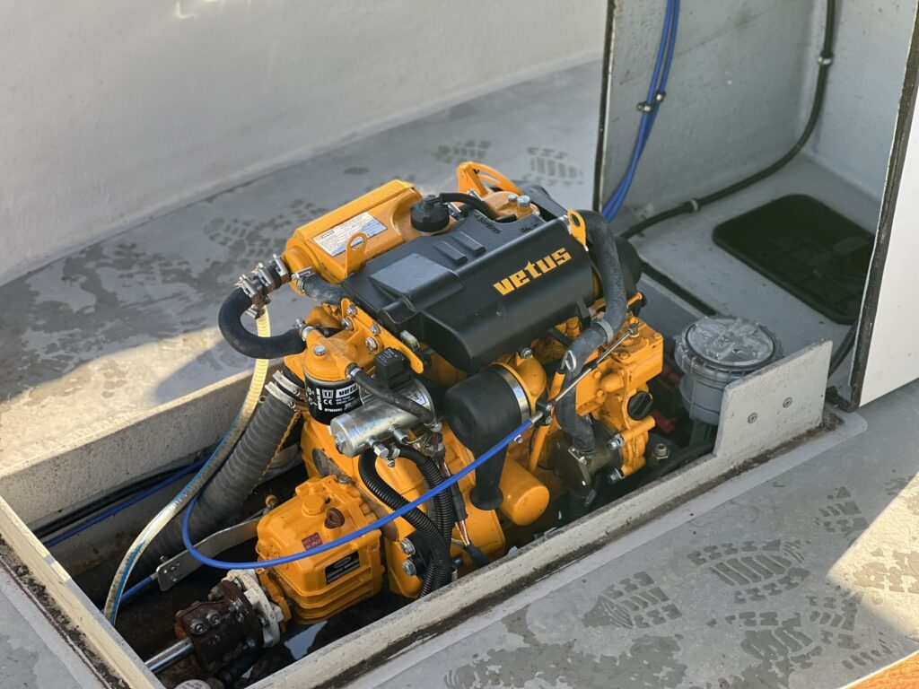 A Vetus marine engine in a compartment, featuring an orange and black color scheme. The engine includes various hoses and fittings, with a gauge visible to the right. The compartment has a gray surface with faint patterns.