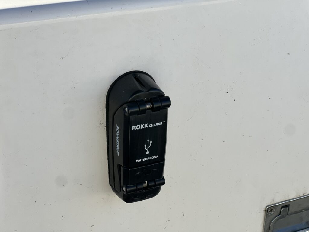 A waterproof charging port marked "ROKK charge" mounted on a white surface. The port has a hinged cover secured to the side, showing a USB symbol.
