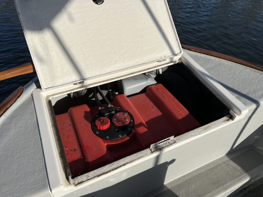 An open compartment on a boat reveals a red fuel tank with a black cap, visible inside a white casing. The surrounding area includes a wooden trim and reflections of light on the water in the background.