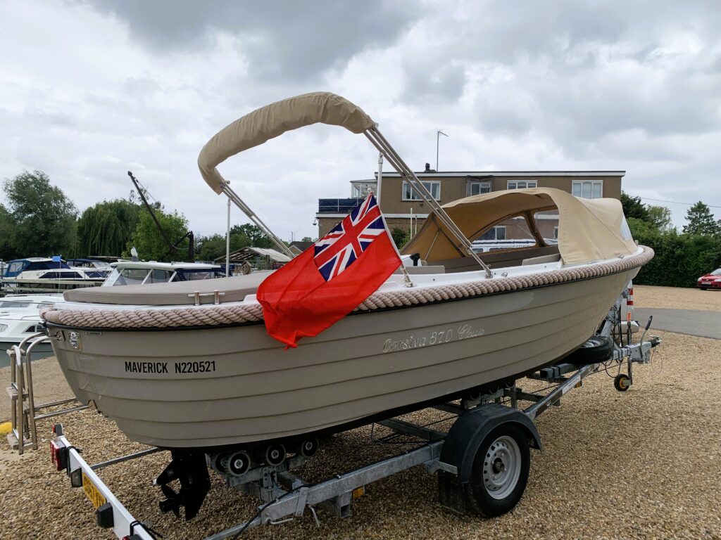 A light brown boat named "Maverick" with registration number N202521 is parked on a trailer. A red flag with the Union Jack is displayed at the stern, and the boat has a beige canopy fitted over its front seating area.