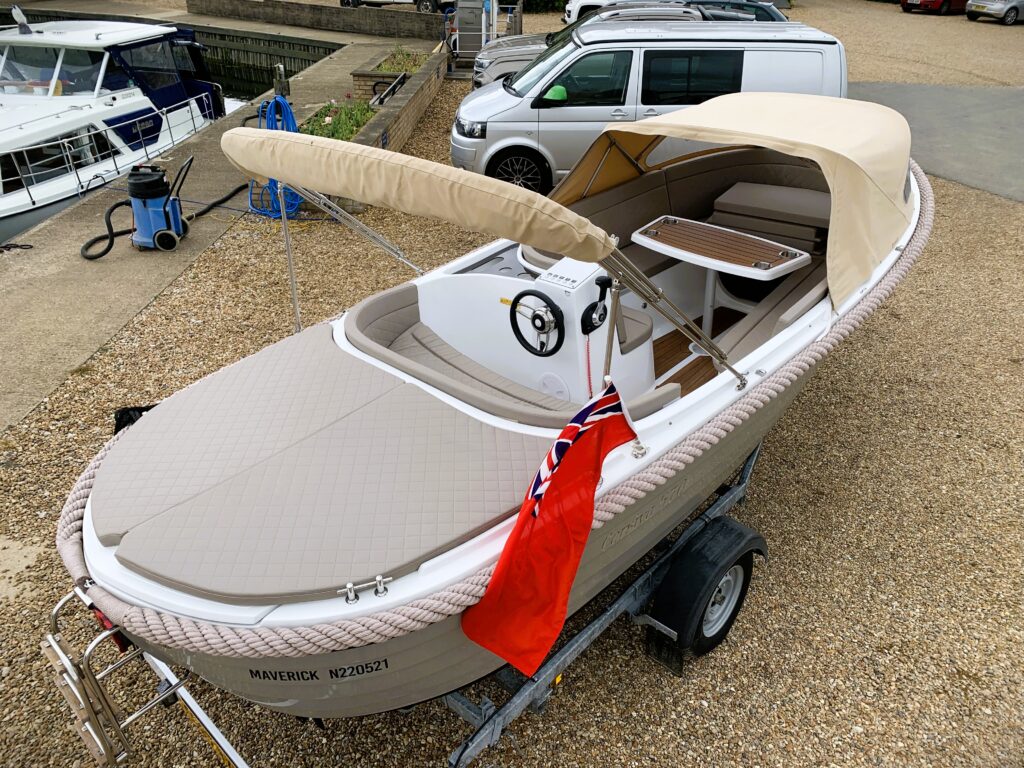 A tan and white boat with a full canopy, resting on a trailer. The boat features an open cockpit with a steering wheel and a seating area. In the background, there are other boats and vehicles in a parking area.