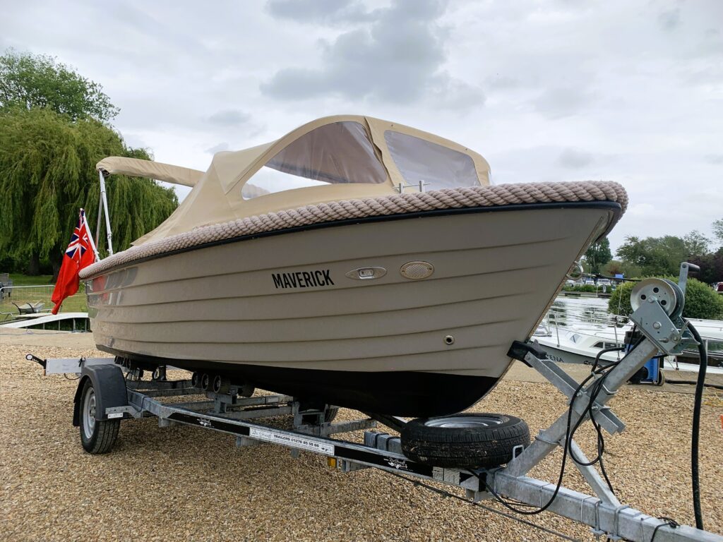 A light beige boat named 'Maverick' is displayed on a trailer. It features a tan bimini top and is parked on a gravel surface, surrounded by greenery and watercraft in the background.