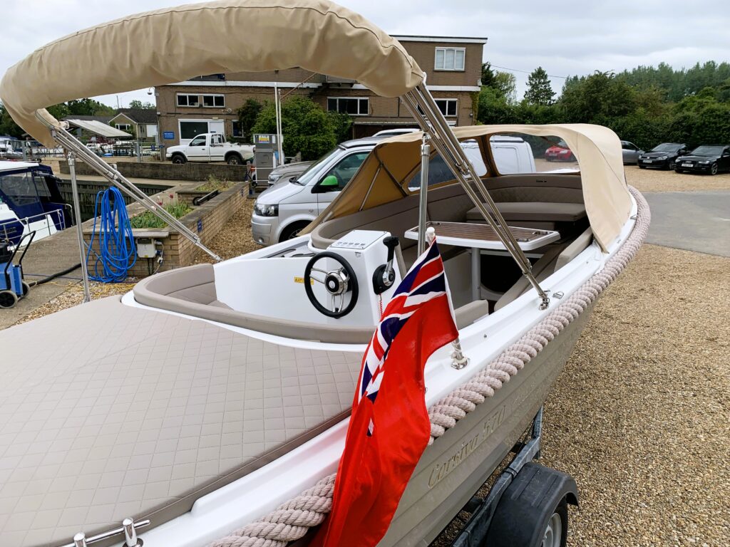 A Corsiva 570 Classic boat on a trailer, featuring a plush tan canopy and a white interior. A Union Jack flag is displayed at the stern, and the background includes parked cars and greenery.