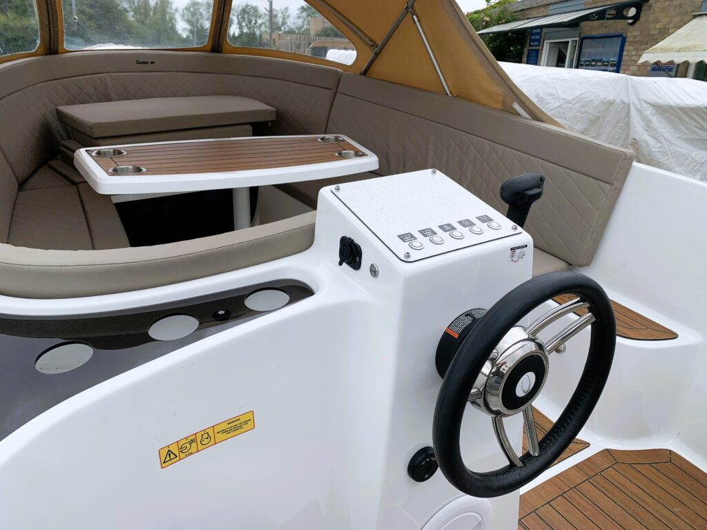 The interior of a Corsiva 570 Classic boat features a clean white dashboard with controls and a black steering wheel. Light brown wood accents are seen on the floor and table, while cushioned seating surrounds the area, providing a spacious and comfortable layout.